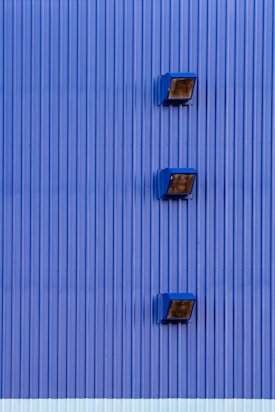Three blue industrial-style lights are mounted on a vertically corrugated blue metal wall, with evenly spaced intervals. The wall texture is uniform with a modern and sleek appearance.