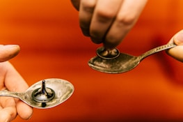 Two hands hold metallic spoons, each containing a spinning top. The background is a warm, reddish-orange color, creating a contrast with the shiny silver surface of the spoons and the reflective tops.