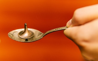 A small metal spinning top is placed on a silver spoon held by a hand. The background is a warm, vivid orange, creating a striking contrast with the metallic objects.