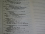 A close-up view of a page from a book containing text. The text appears to be poetic, with numbered lines. The background is blurred, emphasizing the text on the page. The text is black on a light background, and there is a margin with dotted lines on the right side of the page.