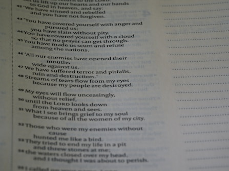 A close-up view of a page from a book containing text. The text appears to be poetic, with numbered lines. The background is blurred, emphasizing the text on the page. The text is black on a light background, and there is a margin with dotted lines on the right side of the page.