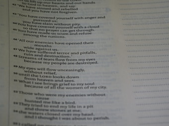 A close-up view of a page from a book containing text. The text appears to be poetic, with numbered lines. The background is blurred, emphasizing the text on the page. The text is black on a light background, and there is a margin with dotted lines on the right side of the page.