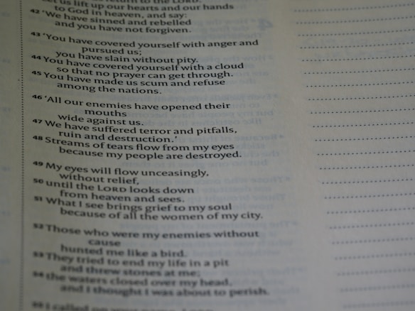 A close-up view of a page from a book containing text. The text appears to be poetic, with numbered lines. The background is blurred, emphasizing the text on the page. The text is black on a light background, and there is a margin with dotted lines on the right side of the page.