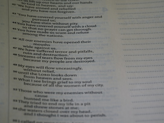 A close-up view of a page from a book containing text. The text appears to be poetic, with numbered lines. The background is blurred, emphasizing the text on the page. The text is black on a light background, and there is a margin with dotted lines on the right side of the page.