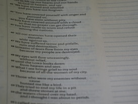 A close-up view of a page from a book containing text. The text appears to be poetic, with numbered lines. The background is blurred, emphasizing the text on the page. The text is black on a light background, and there is a margin with dotted lines on the right side of the page.