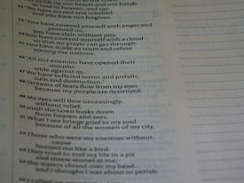 A close-up view of a page from a book containing text. The text appears to be poetic, with numbered lines. The background is blurred, emphasizing the text on the page. The text is black on a light background, and there is a margin with dotted lines on the right side of the page.