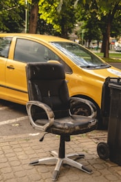 A worn-out black office chair is placed on a paved sidewalk. It is missing parts and shows signs of heavy use. Behind it, a yellow car is parked, partially visible with a lush green park in the background. A trash bin is also seen beside the chair.