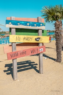A colorful wooden sign on a sandy beach with four horizontal planks painted in different colors. The top plank is blue with painted umbrellas, the second is green, the third is yellow with the text 'Nu AVEM wifi', and the bottom is red with the text 'Vorbiți între voi!' and cartoon eyes. A palm tree and a clear blue sky are visible in the background.
