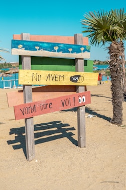 A colorful wooden sign on a sandy beach with four horizontal planks painted in different colors. The top plank is blue with painted umbrellas, the second is green, the third is yellow with the text 'Nu AVEM wifi', and the bottom is red with the text 'Vorbiți între voi!' and cartoon eyes. A palm tree and a clear blue sky are visible in the background.