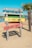 A colorful wooden sign on a sandy beach with four horizontal planks painted in different colors. The top plank is blue with painted umbrellas, the second is green, the third is yellow with the text 'Nu AVEM wifi', and the bottom is red with the text 'Vorbiți între voi!' and cartoon eyes. A palm tree and a clear blue sky are visible in the background.