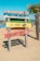 A colorful wooden sign on a sandy beach with four horizontal planks painted in different colors. The top plank is blue with painted umbrellas, the second is green, the third is yellow with the text 'Nu AVEM wifi', and the bottom is red with the text 'Vorbiți între voi!' and cartoon eyes. A palm tree and a clear blue sky are visible in the background.