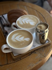 Two cups of coffee with intricate latte art placed on a wooden table alongside a sugar jar and some croissants on a tray. The scene is set in a cozy, inviting atmosphere, perfect for a casual breakfast or coffee break.