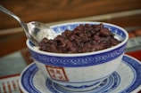 A bowl filled with cooked red beans is placed on a matching saucer. The bowl features traditional blue and white patterns, and a spoon rests inside it.