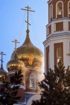 Golden domes of an Orthodox church gleam in the sunlight, adorned with crosses on top. A tall bell tower is visible, with arched windows. Pine trees partially obscure the view, and some mist or smoke can be seen near the base of the domes.