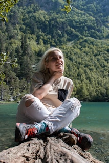 A person with light hair is sitting cross-legged on a log by a clear lake with a lush forested mountain in the background, appearing thoughtful and relaxed. They are wearing a beige shirt with a black pocket, ripped white jeans, colorful socks, and hiking shoes.