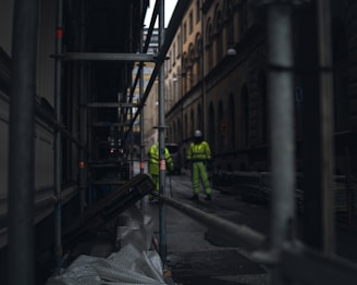 A narrow urban street flanked by scaffolding and tall buildings, with two construction workers in high-visibility clothing and helmets standing in the background. The scene is dimly lit, creating a sense of early morning or late afternoon.