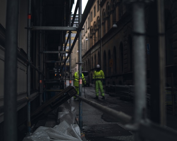 A narrow urban street flanked by scaffolding and tall buildings, with two construction workers in high-visibility clothing and helmets standing in the background. The scene is dimly lit, creating a sense of early morning or late afternoon.