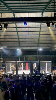 A crowded indoor arena with a cage fight taking place in the center. Audience members surround the cage, with some people involved in organizing the event inside. The arena is well-lit, and the ceiling structure is visible above.