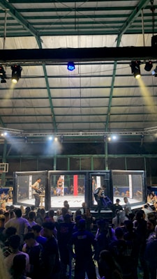 A crowded indoor arena with a cage fight taking place in the center. Audience members surround the cage, with some people involved in organizing the event inside. The arena is well-lit, and the ceiling structure is visible above.