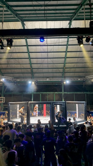 A crowded indoor arena with a cage fight taking place in the center. Audience members surround the cage, with some people involved in organizing the event inside. The arena is well-lit, and the ceiling structure is visible above.