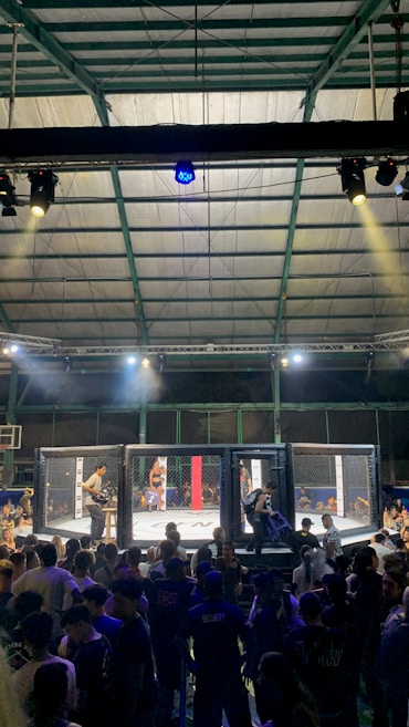 A crowded indoor arena with a cage fight taking place in the center. Audience members surround the cage, with some people involved in organizing the event inside. The arena is well-lit, and the ceiling structure is visible above.