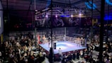 A crowded indoor arena with a boxing ring at the center, surrounded by an audience seated at tables. The scene is illuminated by overhead lights, casting a vibrant glow over the ring where two fighters are preparing for a match. Spectators are engaged, capturing the event with cameras and enjoying the lively atmosphere.