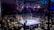 A crowded indoor arena with a boxing ring at the center, surrounded by an audience seated at tables. The scene is illuminated by overhead lights, casting a vibrant glow over the ring where two fighters are preparing for a match. Spectators are engaged, capturing the event with cameras and enjoying the lively atmosphere.