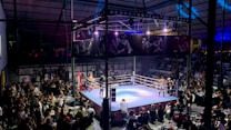 A crowded indoor arena with a boxing ring at the center, surrounded by an audience seated at tables. The scene is illuminated by overhead lights, casting a vibrant glow over the ring where two fighters are preparing for a match. Spectators are engaged, capturing the event with cameras and enjoying the lively atmosphere.