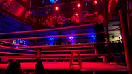 Wide angle of a nighttime outdoor boxing ring, bathed in moody neon lighting with a fighter in stance.