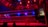 An empty boxing ring is illuminated by dramatic red and blue spotlights inside a dimly-lit arena. A small stool is placed in the center of the ring, surrounded by ropes. The arena is adorned with banners depicting action scenes, and a few silhouettes of spectators can be seen in the background.