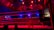 An empty boxing ring is illuminated by dramatic red and blue spotlights inside a dimly-lit arena. A small stool is placed in the center of the ring, surrounded by ropes. The arena is adorned with banners depicting action scenes, and a few silhouettes of spectators can be seen in the background.