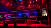 An empty boxing ring is illuminated by dramatic red and blue spotlights inside a dimly-lit arena. A small stool is placed in the center of the ring, surrounded by ropes. The arena is adorned with banners depicting action scenes, and a few silhouettes of spectators can be seen in the background.