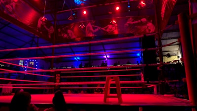 An empty boxing ring is illuminated by dramatic red and blue spotlights inside a dimly-lit arena. A small stool is placed in the center of the ring, surrounded by ropes. The arena is adorned with banners depicting action scenes, and a few silhouettes of spectators can be seen in the background.
