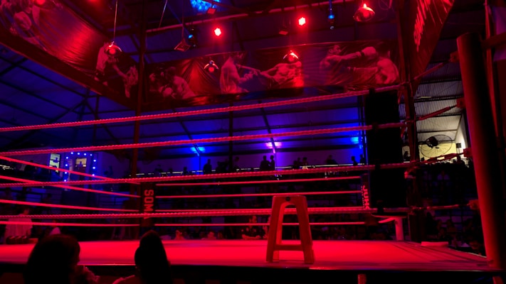 An empty boxing ring is illuminated by dramatic red and blue spotlights inside a dimly-lit arena. A small stool is placed in the center of the ring, surrounded by ropes. The arena is adorned with banners depicting action scenes, and a few silhouettes of spectators can be seen in the background.