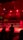 A wrestling ring is illuminated with red lighting, creating a dramatic atmosphere. Several people are visible around the ring, with silhouetted spectators in the foreground. Banners or art adorn the upper walls, and the venue has an industrial ceiling with exposed beams.
