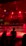 A wrestling ring is illuminated with red lighting, creating a dramatic atmosphere. Several people are visible around the ring, with silhouetted spectators in the foreground. Banners or art adorn the upper walls, and the venue has an industrial ceiling with exposed beams.