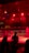 A wrestling ring is illuminated with red lighting, creating a dramatic atmosphere. Several people are visible around the ring, with silhouetted spectators in the foreground. Banners or art adorn the upper walls, and the venue has an industrial ceiling with exposed beams.