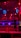 A brightly lit boxing or wrestling ring with vivid red and blue lights. Banners with images of fighters hang from the ceiling. The ropes of the ring are prominently visible, and a stool is placed inside the ring. The arena is filled with an audience, silhouetted against the colorful lighting.
