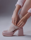 A person wearing a soft pink outfit is putting on a lace sock with floral patterns. The sock is being adjusted on the foot, which is wearing a chunky pink heel. The scene has a soft and delicate aesthetic.