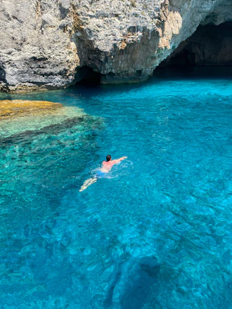 Swimmer cutting through vibrant turquoise ocean waves near rocky coastal landmarks under a bright sky.