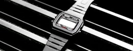 A black digital wristwatch with a rectangular face lies across a series of parallel metallic bars. The watch displays a simple, classic design with a digital screen. The parallel bars provide a contrasting backdrop, highlighting the watch’s sleek appearance.