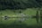 A serene landscape featuring a lush green hillside descending to a calm body of water. Several rustic buildings, including a main cabin and smaller sheds, are situated near the water's edge. The area is surrounded by dense forest with majestic trees. Grazing animals and a few people can be seen at a distance.