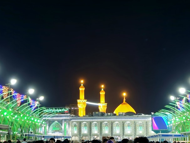 A beautifully illuminated mosque with golden domes and minarets glowing against the night sky. The structure is adorned with green lighting and intricate patterns. The foreground shows a crowd of people, and colorful lights decorate the surrounding area, creating a vibrant and festive atmosphere.