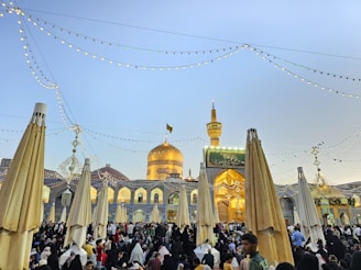 A beautiful courtyard filled with a large gathering of people, featuring a prominent gold-domed mosque in the background. Decorative lights are strung overhead, adding a festive atmosphere. The architecture includes arches and intricate tile work.