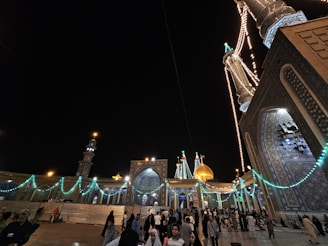 A nighttime scene depicting a grand mosque with illuminated minarets and domes adorned with intricate tile work. Strings of lights illuminate the architectural features, creating a festive atmosphere. A large gathering of people is present, contributing to a lively and vibrant environment.