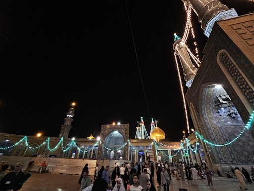 A nighttime scene depicting a grand mosque with illuminated minarets and domes adorned with intricate tile work. Strings of lights illuminate the architectural features, creating a festive atmosphere. A large gathering of people is present, contributing to a lively and vibrant environment.