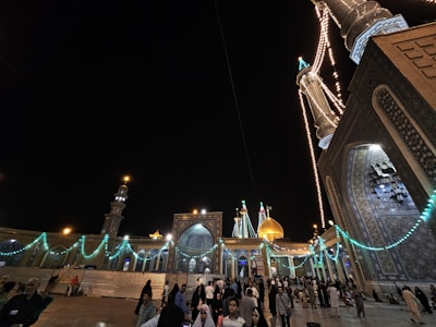 A nighttime scene depicting a grand mosque with illuminated minarets and domes adorned with intricate tile work. Strings of lights illuminate the architectural features, creating a festive atmosphere. A large gathering of people is present, contributing to a lively and vibrant environment.