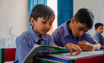 Two young boys wearing blue checkered school uniforms are sitting at a desk in a classroom, focused on their studies. One boy is reading from a colorful book while the other is writing diligently in a notebook. The classroom has a calm atmosphere with a blue door and simple, light-colored walls in the background.