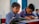 Two young boys wearing blue checkered school uniforms are sitting at a desk in a classroom, focused on their studies. One boy is reading from a colorful book while the other is writing diligently in a notebook. The classroom has a calm atmosphere with a blue door and simple, light-colored walls in the background.