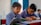 Two young boys wearing blue checkered school uniforms are sitting at a desk in a classroom, focused on their studies. One boy is reading from a colorful book while the other is writing diligently in a notebook. The classroom has a calm atmosphere with a blue door and simple, light-colored walls in the background.